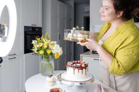 Online Gifting - Side view of positive young female food blogger in casual clothes and apron introducing yummy cakes and smiling while standing in front of ring lamp and recording vlog on smartphone