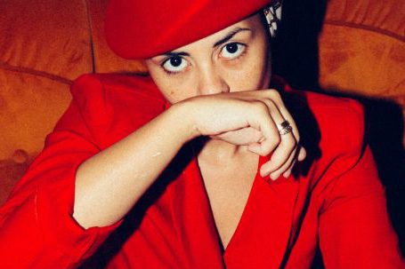 Fashion Fascination - From above of determined female model wearing trendy suit and retro hat sitting in comfortable armchair and covering mouth with hand while looking at camera