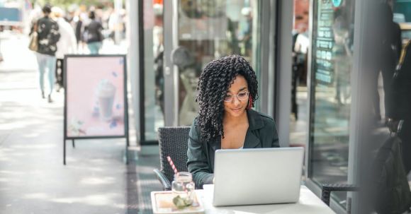 Responsible Online Shopping - High angle of pensive African American female freelancer in glasses and casual clothes focusing on screen and interacting with netbook while sitting at table with glass of yummy drink on cafe terrace in sunny day
