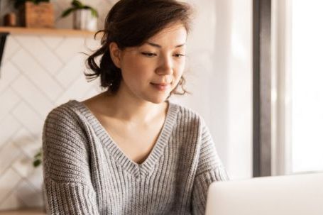 Online Appliance Shopping. - Young female in gray sweater sitting at wooden desk with laptop and bottle of milk near white bowl while browsing internet on laptop during free time at home